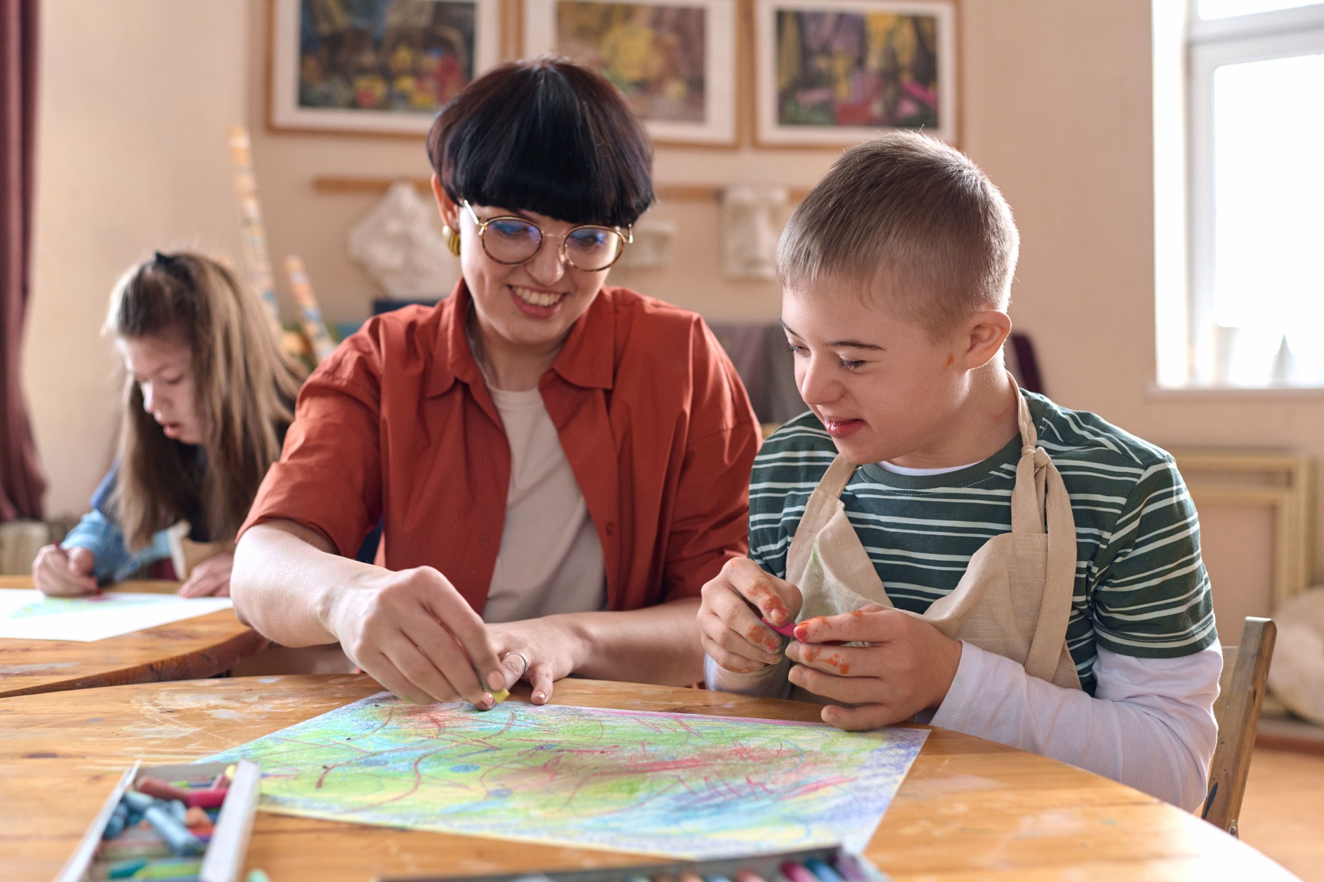 Smiling Boy with Disability Enjoying Art Class in Studio with Mentor Assisting